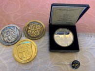 Auf einer beigen Tischdecke mit Schnörkeln liegen drei Medaillen in gold, silber und branze mit dem Kiedricher Wappen mit der Burgruine Scharfenstein und dem mit einem Kreuz verbundenem Doppelrad. ebenso liegt in einer schwarzen aufgeklappten Medaillendose eine silberne Medaile mit  der Beschriftung Kiedrich und Rheingau und den Kiedricher Wahrzeichen. Davor liegt eine schwarz-silberne Anstecknadel.