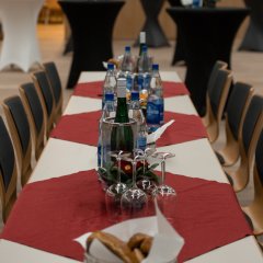 Eine lange Tafel, eingedeckt mit weinroten kleinen viereckigen Tischdecken, auf denen Brotkörbe mit Laugengebäck, Wasserflaschen, Weinflaschen, Weinkühler und Weingläser stehen. Im Hintergrund sind eingedeckte Stehtische mit Hussen.