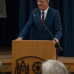 Bürgermeister Steinmacher, in blauem Anzug, mit weißem Hemd, Kravatte und blauer Brille  steht hinter einem Mikrofon an einem Rednerpult und lacht. Am Rednerpult ist das Kiedricher Wappen erkennbar und im Hintergrund ist ein blauer Vorhang.
