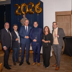 Fünf Männer und eine Frau stehen nebeneinander. Vier davon halten ein gefülltes Sektglas in der Hand. Alle tragen Sakkos  und dunkle Hosen dazu. Die Frau trägt einen schwarzen Hosenanzug. Im Hintergrund sieht man die Zahl 2026 aus vier einzelnen goldenen Folienballons vor einem blauen Bühnenvorhang.