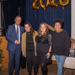 Ein Mann und drei Frauen stehen vor einem blauem Bühnenvorhang, vor dem man die Zahl 2026 aus vier einzelnen goldenen Folienballons sieht. Der Mann trägt eine Brille, ein weißes Hemd, eine Krawatte und einen blauen Anzug. Die Frauen sind dunkel gekleidet, eine Frau im kurzem Kleid und die anderen mit Hosen und langärmelig.