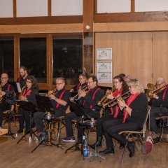 Eine Bank, bestehend aus dreizehn Personen sitzen auf Holzstühlen.Sie tragen schwarze Hosen und Hemden bzw. Blusen und die Männer tragen rote Hosenträge und die Frauen rote Schals. Es sind Blasinstrumente (Trompeten und Saxophone) und ein Schlagzeug zu erkennen.