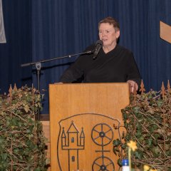 Neben einem Rednerpult aus Holz, auf dem aus Metall das Kiedricher Wappen, d.h. die Burgruine Scharfenstein und das Doppelrad erkennbar ist, stehen zwei Efeu-Türme. Hinter dem Rednerpult steht eine Frau mit kurzen Haaren und einem schwarzem Oberteil und spricht in ein dunkles Mikrofon. Im Hintergrund ist ein blauer Bühnenvorhang.