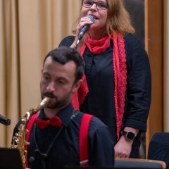 Ein Mann sitzt und spielt Saxophon. Er trägt ein schwarzes Hemd und rote Hosenträger sowie eine rote Fliege. Dahinter steht eine Frau in einer schwarzen Bluse und einem roten Schal und einer scharzen Brille. Sie hat schuterlange braune Haare und hält ein Mikrofon an ihren Mund und singt.