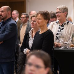 Mehrere Personen stehen in einem Saal neben einem Stehtisch, auf dem leere Sektgläser und Laugengebäck stehen. 