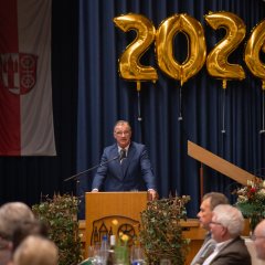 Ein Mann mit weißem Hemd, dunkelblauem Anzug und blauer Krawatte steht hinter einem Rednerpult aus Holz. Neben einem Rednerpult, auf dem aus Metall das Kiedricher Wappen, d.h. die Burgruine Scharfenstein und das Doppelrad erkennbar ist, stehen zwei Efeu-Türme. Im Hintergrund ist ein blauer Bühnenvorhang und davor sieht man die Zahl 2026 aus vier einzelnen goldenen Folienballons. Ebenso hängt dort in rot/weiß die Kiedricher Flagge.