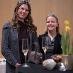 Hinter einem Bistrotisch mit dunkler Husse und einer Osterglocke als Blumendeko, stehen zwei Frauen mit blonden, langen, offenen Haaren und Kronen auf dem Kopf. Die linke Frau mit Brille trägt ein schwarzes Oberteil und eine graue offene Strickjacke und rechts die Frau trägt einen schwarzen Blazer und darunter ein graues Oberteil mit Nadelstreifen.