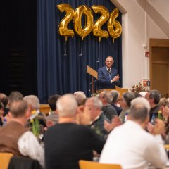 Ein Mann mit weißem Hemd, dunkelblauem Anzug und blauer Krawatte steht hinter einem Rednerpult aus Holz. Im Hintergrund ist ein blauer Bühnenvorhang und davor sieht man die Zahl 2026 aus vier einzelnen goldenen Folienballons. Im Vordergrund sitzen mehrere Personen an langen Tischreihen.
