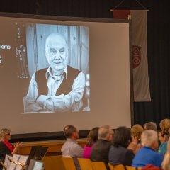 Vor einem blauen Bühnenvorhang steht eine große Leinwand, auf die ein Bild in schwarz-weiß projiziert ist. Auf dem Bild ist ein Mann ohne Haare mit hellem Hemd und schwarzem Pullunder und verschränkten Armen, der lacht. Vor der Bühne sitzen Personen an langen Tischen und links sitzen Personen einer Band, in schwarz und rot gekleidet mit ihren Blasinstrumenten und Notenständern.