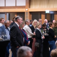 Mehrere Personen stehen in einem Saal hinter einem Stehtisch, auf dem leere Sektgläser und Laugengebäck stehen. 