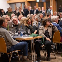 Mehrere Personen sitzen an langen Tischen die  mit grauen viereckigen kleinen Tischdecken und mehrere Osterglocken in Töpfen dekoriert sind. Im Hintergrund stehen weitere Personen in dem Saal.