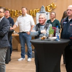 Mehrere Personen stehen in einem Saal hinter und neben einem Stehtisch, auf dem Sektgläser, eine Wasserflasche und Laugengebäck stehen.