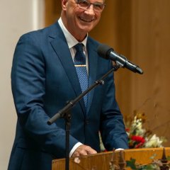 Ein Mann mit blauer Brille, weißem Hemd, dunkelblauem Anzug und blauer Krawatte steht hinter einem Rednerpult aus Holz und lacht. Danebn steht ein Mikrofon mit Ständer und eine Efeu-Pflanze und rechts im Hintergrund ist ein rot-weißer Blumenstrauß. 