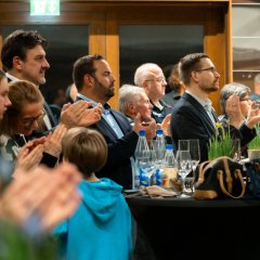 Mehrere Personen stehen in einem Saal neben und hinter zwei Stehtisch, auf denen leere Sektgläser, Osterglocken in Töpfen, Wasserflaschen und Laugengebäck stehen und applaudieren. 