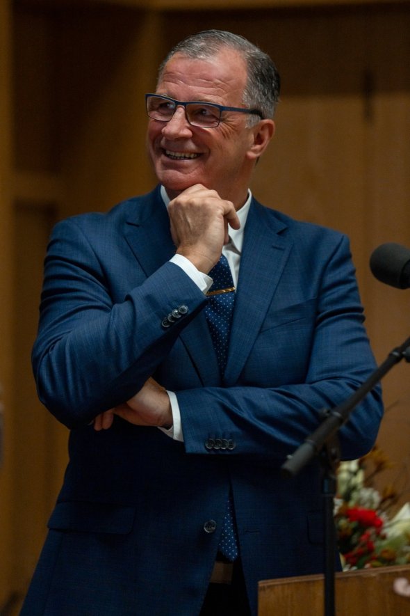 Ein Mann mit blauer Brille, weißem Hemd, dunkelblauem Anzug und blauer Krawatte steht hinter einem Rednerpult aus Holz, hat die Arme vor dem Körper verschränkt und eine Hand ist am Kinn. Er lacht. Daneben steht ein Mikrofon und im Hintergrund ist ein rot-weißer Blumenstrauß. 