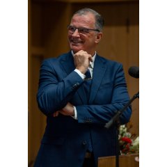 Ein Mann mit blauer Brille, weißem Hemd, dunkelblauem Anzug und blauer Krawatte steht hinter einem Rednerpult aus Holz, hat die Arme vor dem Körper verschränkt und eine Hand ist am Kinn. Er lacht. Daneben steht ein Mikrofon und im Hintergrund ist ein rot-weißer Blumenstrauß. 