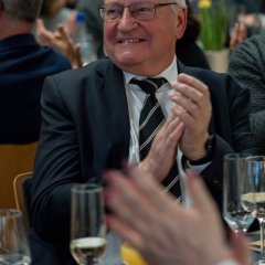 An einem Tisch, auf dem volle und leere Sektgläser stehen, sitzt ein Mann mit Brille, weißem Haar, einem weißen Hemd, einer schwarzen Krawatte mit weißen Streifen und einem schwarzem Sakko und applaudiert.