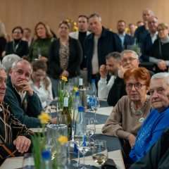 Mehrere Personen sitzen an einem langen Tisch der mit grauen viereckigen kleinen Tischdecken und mehrere Osterglocken in Töpfen dekoriert ist. Im Hintergrund stehen weitere Personen in dem Saal.