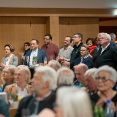 Mehrere Personen stehen in einem Saal. Vor den stehenden Personen sitzen mehrere Personen. 