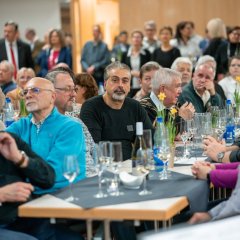 Mehrere Personen sitzen an einem langen Tisch der mit grauen viereckigen kleinen Tischdecken und mehrere Osterglocken in Töpfen dekoriert ist. Im Hintergrund stehen weitere Personen in dem Saal.