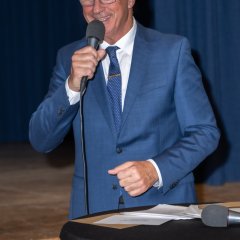 Ein Mann mit blauer Brille, weißem Hemd, dunkelblauem Anzug und blauer Krawatte steht hinter einem Stehtisch mit dunkler Husse und hält ein Mikrofon in der Hand, an seinen Mund.