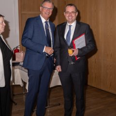 Zwei Männer mit Brille und dunklem Anzug und blauer Krawatte und weißem Hemd. Ein Mann hat einen roten Schnellhefter mit weißen Zetteln drin unter den Arm geklemmt und hat ein Sektglas mit Orangensaft in der Hand. 