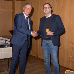 Zwei Männer mit Brille geben sich die Hand. Ein Mann trägt dunklem Anzug, blaue Krawatte und ein weißes Hemd. Der andere Mann trägt eine Jeans, ein blaues Sakko mit weißem Hemd und einen blauen Pullover. Er hat ein Sektglas mit Orangensaft in der Hand. 