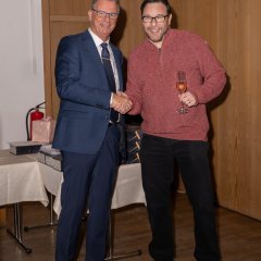 Zwei Männer mit Brille geben sich die Hand. Ein Mann trägt einen dunkelblauen Anzug, eine blaue Krawatte und ein weißes Hemd. Der andere Mann hat eine dunkle Hose und einen rot-melierten Pulover an. Er hat ein gefülltes Sektglas in der Hand. 