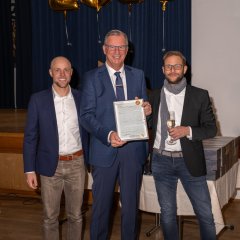 Drei Männer, zwei mit Brille stehen nebeneinander. Alle tragen ein weißes Hemd und dunkle Sakkos. Der Mann in der Mitte trägt eine dunkelblaue Krawatte und hält einen Bilderrahmen in der Hand. Der Mann rechts trägt einen grauen Schal und hat ein gefülltes Sektglas in der Hand. Im Hintergrund sieht man die Zahl 2026 aus vier einzelnen goldenen Folienballons vor einem blauen Bühnenvorhang.