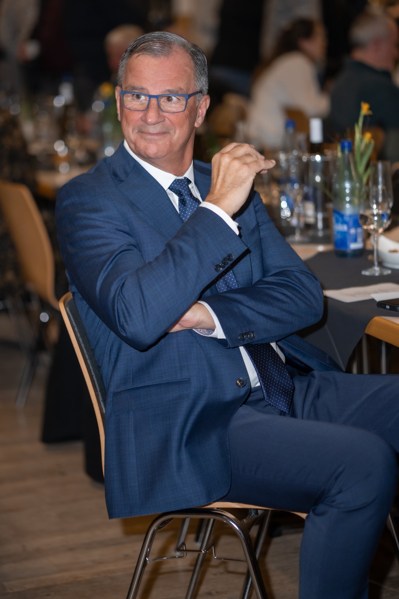 Auf einem Holzstuhl sitzt ein Mann mit Brille, weißem Hemd, blauer Krawatte und blauem Anzug. Er hat beide Arme vor dem Oberkörper und ein Arm reicht nach oben richtung Gesicht. Im Hintergrund ist ein mit Blumen, Gläsern und Flaschen gedeckter Tisch mit grauen Tischdecken erkennbar.