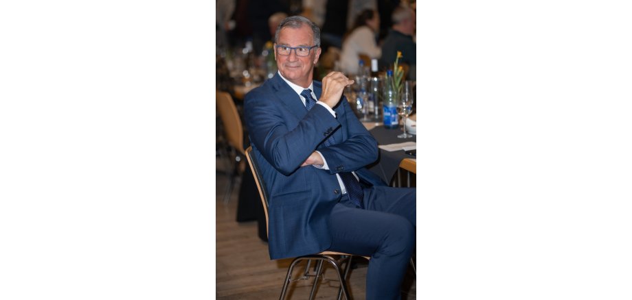 Auf einem Holzstuhl sitzt ein Mann mit Brille, weißem Hemd, blauer Krawatte und blauem Anzug. Er hat beide Arme vor dem Oberkörper und ein Arm reicht nach oben richtung Gesicht. Im Hintergrund ist ein mit Blumen, Gläsern und Flaschen gedeckter Tisch mit grauen Tischdecken erkennbar.