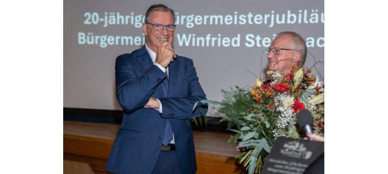 Vor einer Holzbühne, auf der sich eine Leinwand  befindet, stehen zwei Männer, beide mit Brille und Anzug. Der rechte Mann hält einen großen weiß-roten Blumenstrauß in der Hand. 