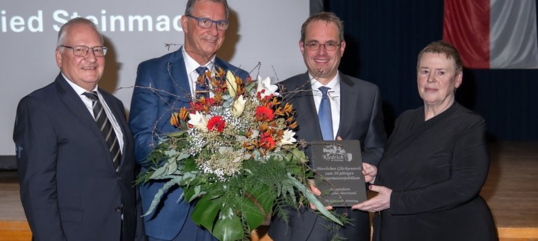 Vor einer Holzbühne, auf der eine Leinwand steht, auf die Bilder und Texte projiziert werden, stehen drei Männer und eine FrauDie Frau Trägt schwarz und hält eine Schieferplatte in der Hand. Die Männer tragen weiße Hemden, brillen, Krawatten und dunkle Anzüge. Der zweite von links hält einen großen weiß-roten Blumenstrauß in der Hand. Vor dem blauen Bühnenvorhang hängt die Kiedricher Flagge in rot-weiß mit der Burgruine Scharfenstein und dem Doppelrad.