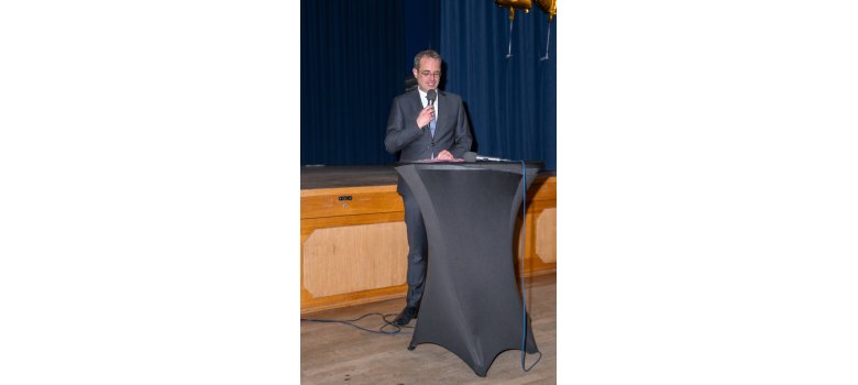 Vor einer Holzbühne mit blauem Vorhang steht ein Mann mit Brille, weißem Hemd, Krawatte und Anzug hinter einem Stehtisch mit grauer Husse und hält ein Mikrofon in der Hand.