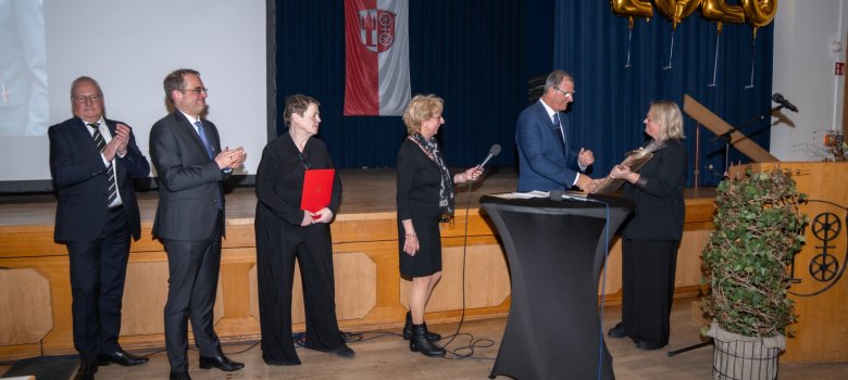 Vor einer Holzbühne, auf der eine Leinwand steht, auf die Bilder projiziert werden, stehen drei Frauen und 3 Männer. Davor steht ein Stehtisch mit grauer Husse und ein Rednerpult aus Holz, vor dem eine Pflanze steht. Vor dem blauen Bühnenvorhang hängt mit goldenen Luftballons die Zahl 2026 und die Kiedricher Flagge in rot-weiß mit der Burgruine Scharfenstein und dem Doppelrad.