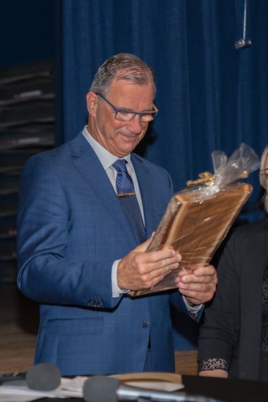 Ein Mann hält eine Holzplatte, die in Klarsichtfolie eingepackt ist, in der Hand. Er trägt eine Brille, ein weißes Hemd, eine blaue Krawatte und einen blauen Anzug. Im Hintergrund ist ein blauer Vorhang erkennbar.