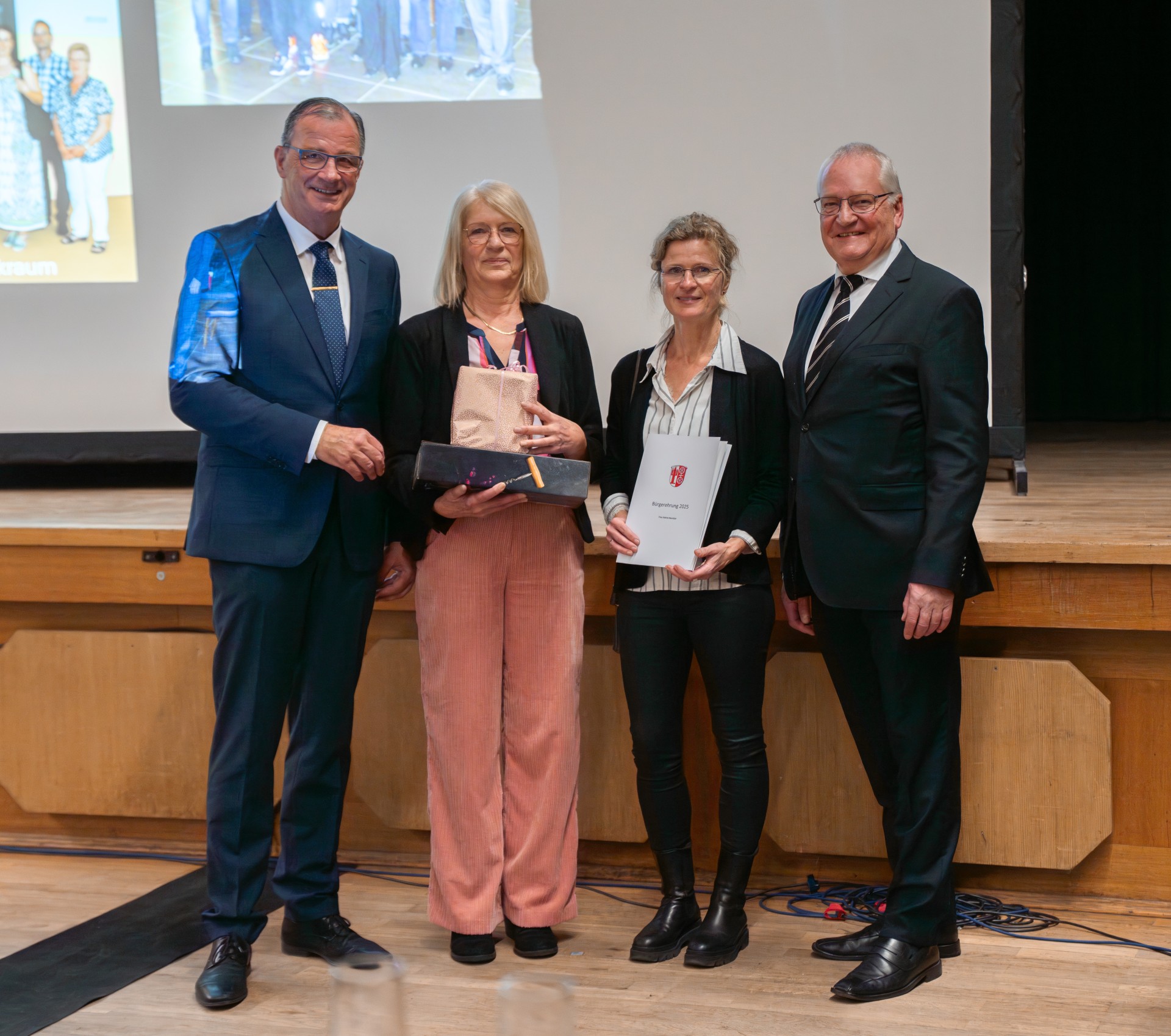 Vor einer Holzbühne, auf der eine Leinwand steht, auf die Bilder projiziert werden, stehen zwei Frauen und links und rechts davon jeweils ein Mann. Die Männer tragen Anzug und die Frauen Blazer. Eine Frau hält eine weiße Urkundenmappe in der Hand.