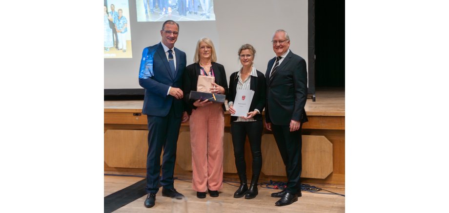 Vor einer Holzbühne, auf der eine Leinwand steht, auf die Bilder projiziert werden, stehen zwei Frauen und links und rechts davon jeweils ein Mann. Die Männer tragen Anzug und die Frauen Blazer. Eine Frau hält eine weiße Urkundenmappe in der Hand.