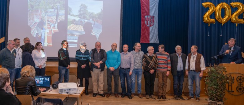 Vor einer Holzbühne, auf der eine Leinwand steht, auf die Bilder projiziert werden, stehen drei Frauen und 12 Männer. An einem Rednerpult steht ein weiterer Mann im Anzug. Vor dem blauen Bühnenvorhang hängt mit goldenen Luftballons die Zahl 202 und die Kiedricher Flagge in rot-weiß mit der Burgruine Scharfenstein und dem Doppelrad.