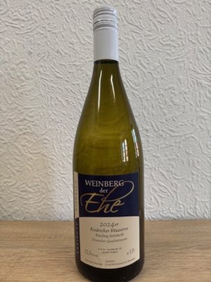 Eine dunkelgrüne Weinflasche mit beige-blauem Etikett mit weißer, schwarzer und goldener Schrift und weißem Aluschrauber, steht vor einer weißen Strukturtapete.