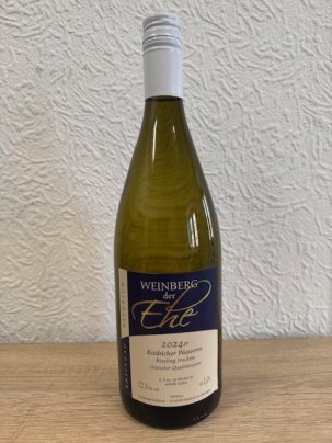 Eine dunkelgrüne Weinflasche mit beige-blauem Etikett mit weißer, schwarzer und goldener Schrift und weißem Aluschrauber, steht vor einer weißen Strukturtapete.