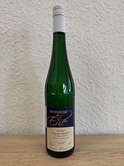 Eine dunkelgrüne Weinflasche, mit Erhebungen/Streifen am Flaschenhals, mit beige-blauem Etikett mit weißer, schwarzer und goldener Schrift und weißem Aluschrauber, steht vor einer weißen Strukturtapete.