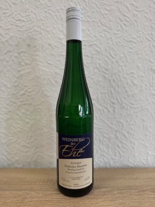 Eine dunkelgrüne Weinflasche, mit Erhebungen/Streifen am Flaschenhals, mit beige-blauem Etikett mit weißer, schwarzer und goldener Schrift und weißem Aluschrauber, steht vor einer weißen Strukturtapete.