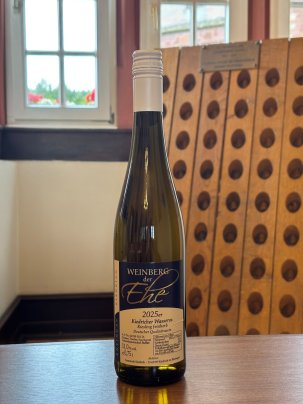 Eine dunkelgrüne Weinflasche, mit beige-blauem Etikett mit weißer, schwarzer und goldener Schrift und weißem Aluschrauber, steht auf einem Holztisch, vor einem Rüttelbrett und weißen Sprossenfenstern, an denen draußen Blumekästen mit grün-roten Blumen hängen.