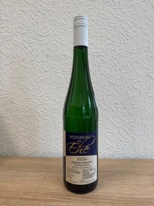 Eine dunkelgrüne Weinflasche, mit Erhebungen/Streifen am Flaschenhals, mit beige-blauem Etikett mit weißer, schwarzer und goldener Schrift und weißem Aluschrauber, steht vor einer weißen Strukturtapete.