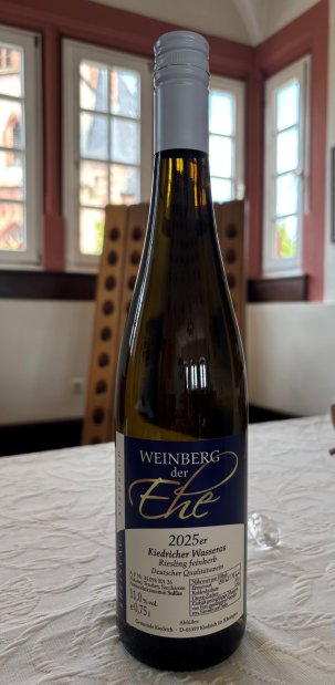 Eine dunkelgrüne Weinflasche, mit beige-blauem Etikett mit weißer, schwarzer und goldener Schrift und weißem Aluschrauber, steht auf einer beigen Strukturtischdecke, vor einem Rüttelbrett und weißen Sprossenfenstern.