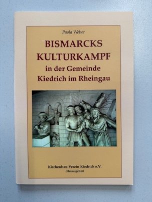 Buchtitel "Bismarcks Kulturkampf in der Gemeinde Kiedrich im Rheingau". Vor einer Mauer mit zwei Rundfenstern steht ein Soldat mit Lanze und Jesus trägt zusammen mit zwei Jüngern das Kreuz. Im Hintergrund sind mehrere Menschen, Häuser und eine Burg zu erkennen. Die Figuren sind aus Stein.