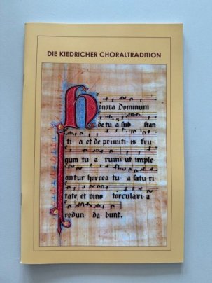 Buchtitel "Die Kiedricher Choraltradition". Auf einem alten Blatt Papier sind Hufnagelnoten und Liedtexte und der erste Buchstabe ist über die ganze Blattseite geschrieben und blau, rot, schwarz und gold verziert.