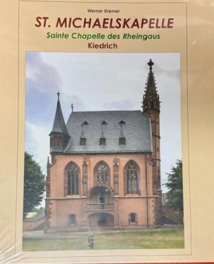 Auf beigem Untergrund ist in Großbuchstaben der Buchtitel "St. Michaelskapelle" und auf dem Bild die Kapelle mit Ihren Türmen, den drei großen Seitenfenstern, den Treppen und dem Balkon dargestellt. Es ist die Wiese, die Bäume und ein bewölkter Himmel zu sehen.