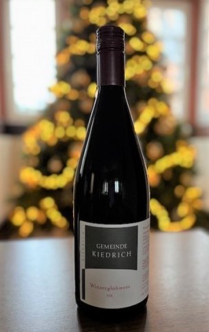 Eine dunkle Weinflasche, mit weiß-grauem Etikett und dunkelrotem Aluschrauber, auf einer braunen Tischplatte. Im Hintergrund ist verschwommen ein Weihnachtsbaum mit leuchtender Lichterkette vor Sprossenfenstern.